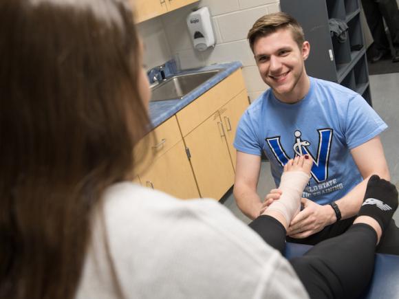 A student majoring in Movement Science. He sits on a stool in front of another student with a bandage wrapped around her ankle. He's studying it as part of his coursework.